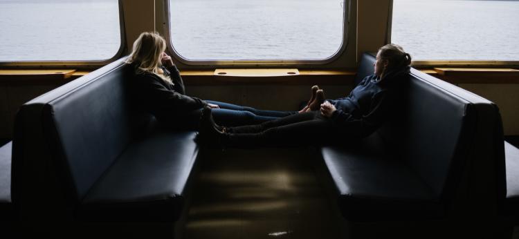Two young women sit in seats looking out the window of a moving ferry.