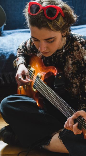 Young woman in bedroom learning how to play an electric guitar looking down to a mobile phone.