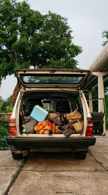 Open car trunk of a white 1990s automobile. The car is packed with a cooler, firewood, and a bunch of bags. Natural warm summer evening light.