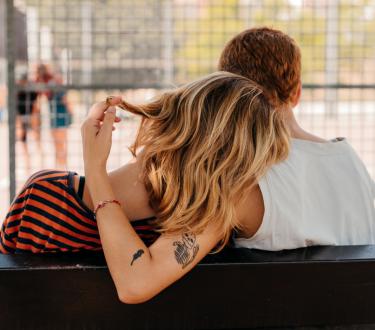 Two young queer women, caressing and showing affection to each other, sitting on a bench at the university campus.
