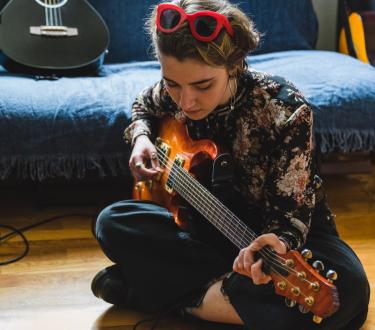 Young woman in bedroom learning how to play an electric guitar looking down to a mobile phone.